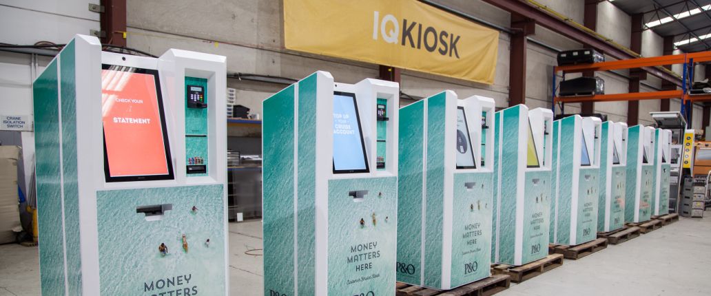 A photo of the IQ KIOSK workshop, with a set of 8 custom cash management kiosks in the foreground and an IQ KIOSK banner in the background